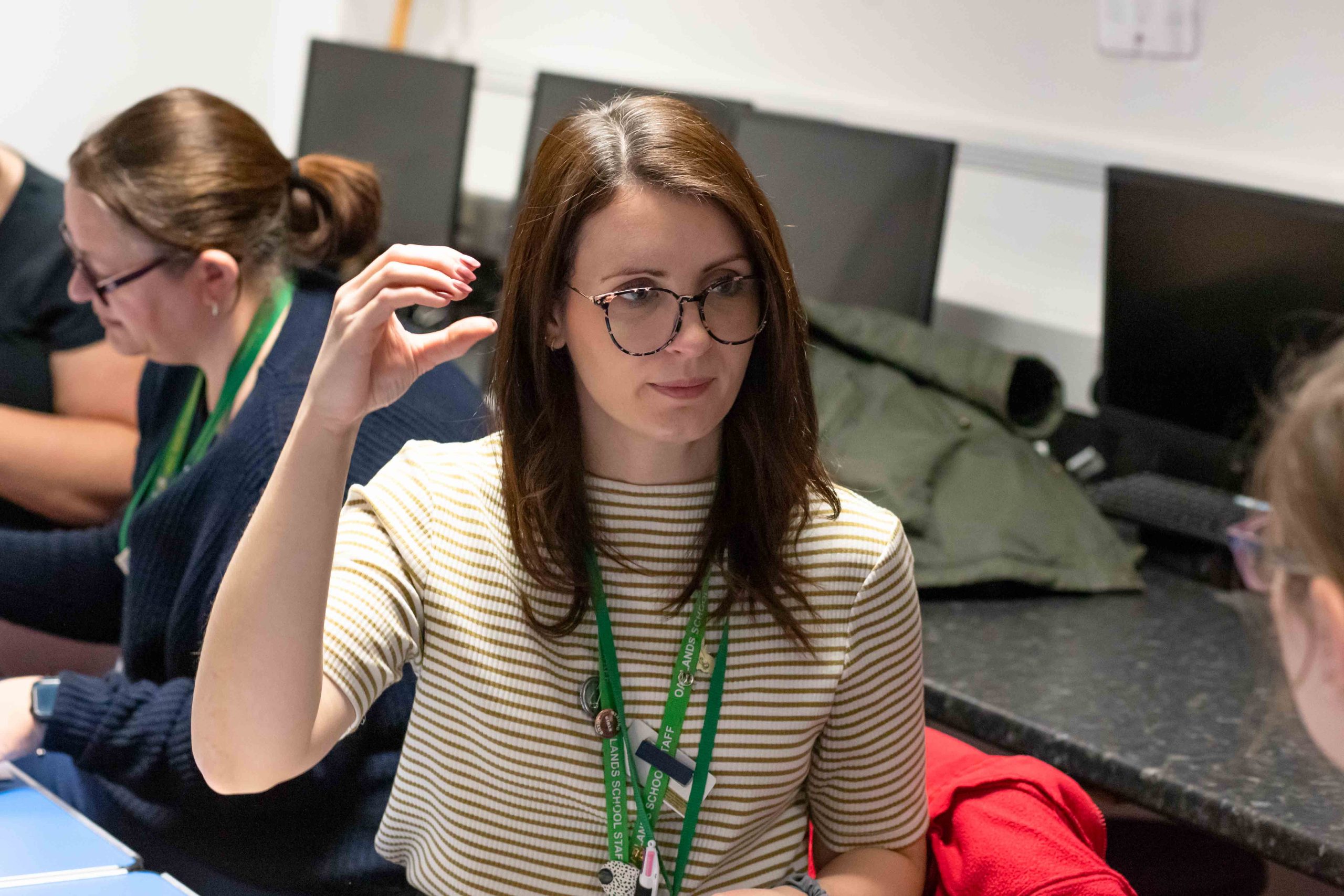 Signalong course participant seated in a classroom, demonstrating a hand sign during a training session. A green staff lanyard is visible, with computer monitors and other course participants seated at desks in the background.